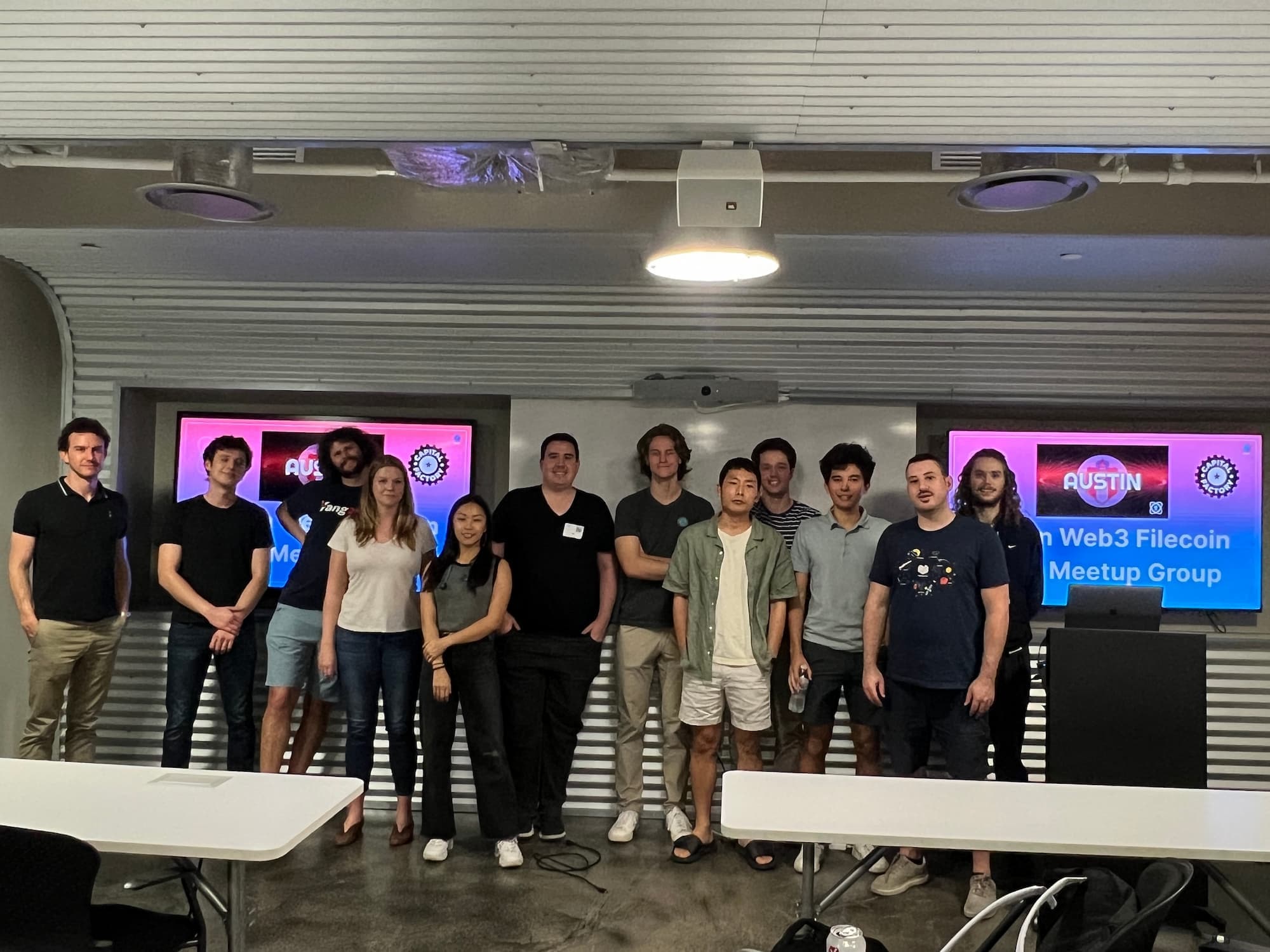 A group of people standing together at the Austin Web3 Filecoin Meetup Group event. They are indoors, and behind them is a screen displaying the event name and logo. The group consists of men and women, casually dressed, smiling and posing for the photo.